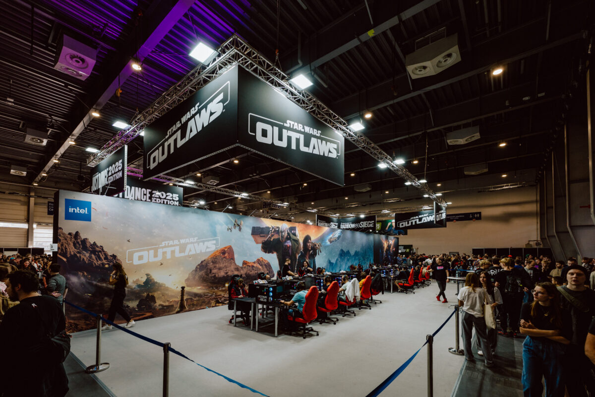 Eine große Spieleveranstaltung mit Menschen, die sich um den 'Star Wars Outlaws'-Stand mit Bannern, Werbegrafiken und Reihen von Computern mit Spielern auf roten Stühlen versammelt haben. Viele Teilnehmer stehen und schauen hinter den Absperrungen zu. 