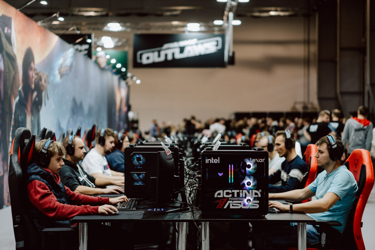 Reihen von Leuten mit Headsets spielen Videospiele auf Desktop-Computern während eines E-Sport-Turniers. Zuschauer stehen im Hintergrund und ein großes Banner mit der Aufschrift 'OUTLAWS' hängt über der Bühne. 
