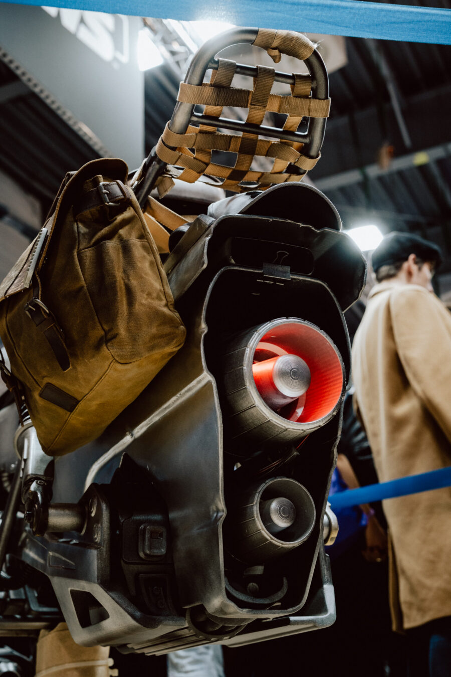 Nahaufnahme eines futuristischen mechanischen Exoskelettbeins mit rot leuchtendem Kern und angebrachtem Zubehör, darunter eine braune Tasche, in einem Raum mit Menschen und blauen Seilbarrieren im Hintergrund.