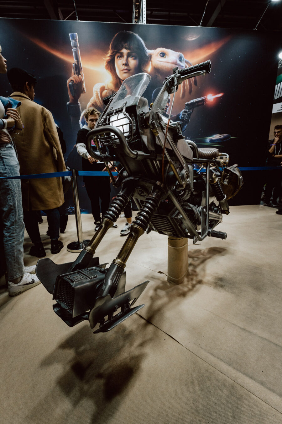 Eine Nachbildung eines Star Wars-Speeder-Bikes steht in einem Raum, um den sich Menschen versammelt haben. Dahinter befindet sich ein großes Poster, auf dem Star Wars-Figuren abgebildet sind, darunter Luke Skywalker und Yoda. 