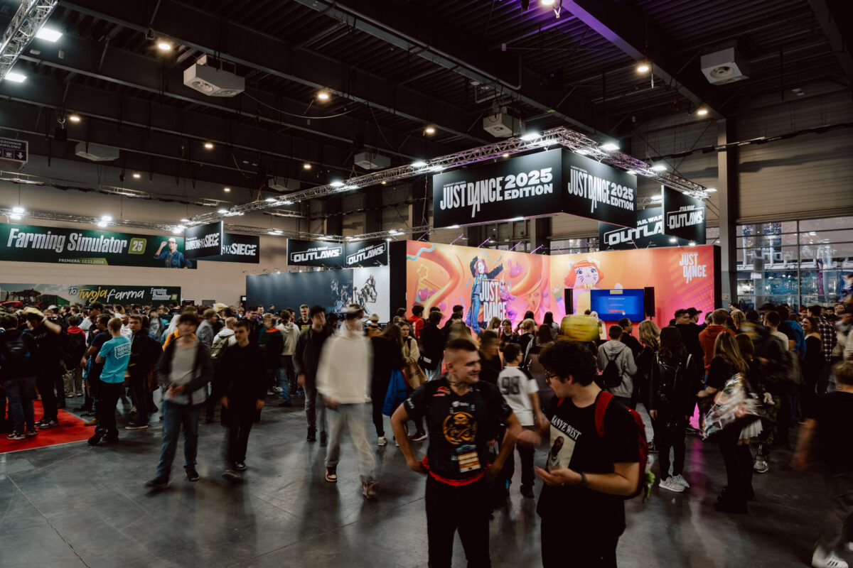 Eine große Menschenmenge versammelt sich auf der Spielemesse. Die Stände von 'Just Dance 2025 Edition' und 'Farming Simulator' sind gut sichtbar. Bunte Auslagen, Banner und Menschen, die sich unterhalten, füllen die geräumige, gut beleuchtete Halle. 