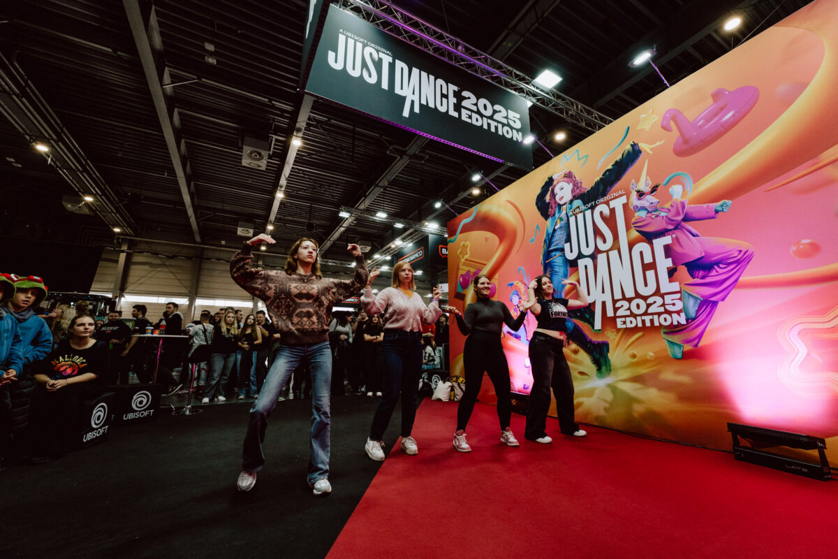 Vier Personen tanzen auf dem roten Teppich vor einer farbenfrohen 'Just Dance 2025 Edition'-Leinwand bei einer Indoor-Veranstaltung, während die Menge zuschaut und große Schilder über ihnen das Spiel bewerben.