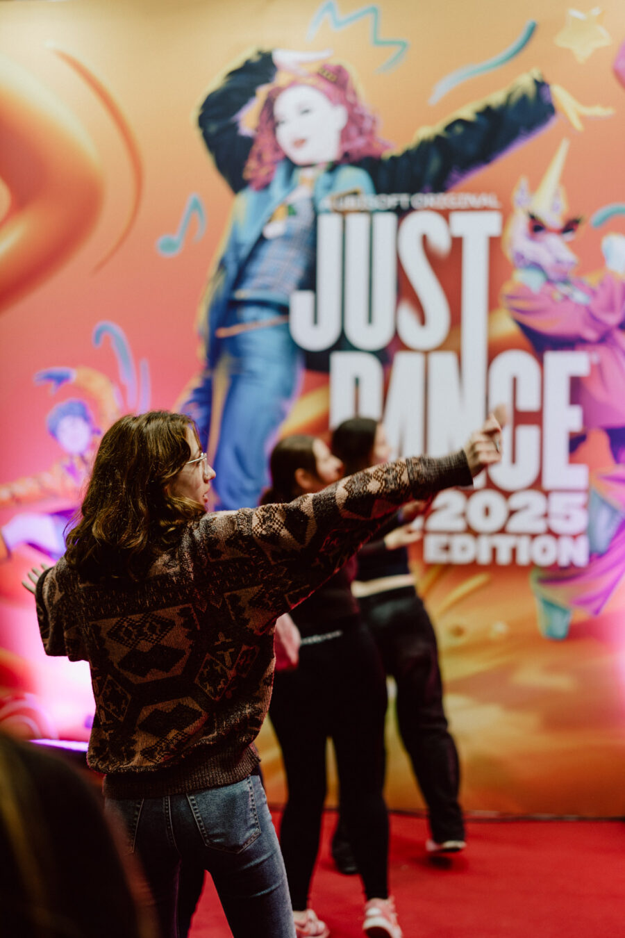 Eine Person in einem gemusterten Pullover tanzt mit ausgestreckten Armen vor einem farbenfrohen 'Just Dance 2025 Edition' Werbehintergrund. Auch andere Personen tanzen im Hintergrund. 