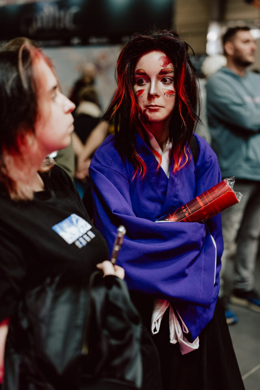 Eine Person in einem violetten Kimono mit dramatischer roter und schwarzer Gesichtsbemalung und einer Perücke hält ein aufgerolltes rotes Plakat. Sie steht neben einer anderen Person in legerer Kleidung bei einer überfüllten Veranstaltung in einer Halle.