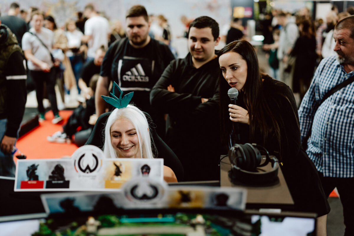 Eine Frau im Cosplay mit weißem Haar und grüner Kopfbedeckung lächelt, während sie an einem Computer sitzt und von Schaulustigen umgeben ist. Eine Frau mit einem Mikrofon spricht neben ihr auf einer belebten Veranstaltung oder einem Kongress. 