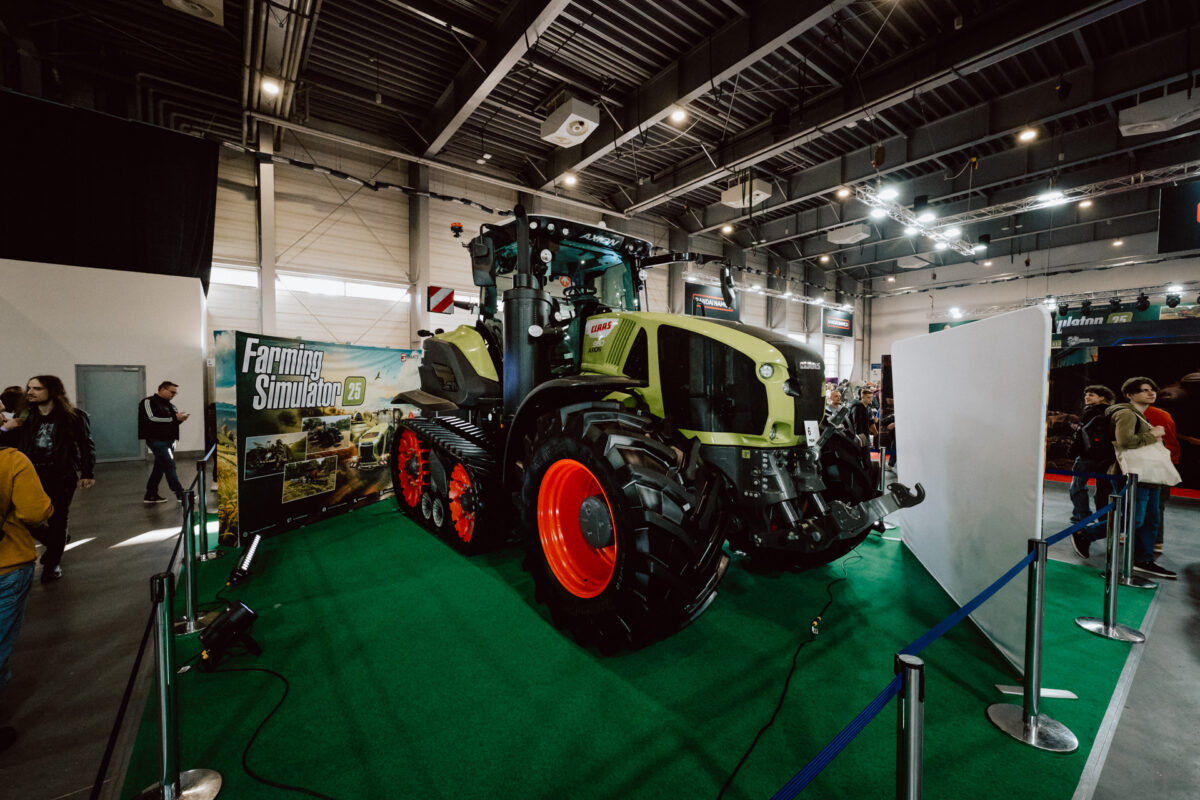 Ein großer grün-schwarzer Traktor mit roten Rädern steht in einem Raum auf einem grünen Teppich, umgeben von blauen Seilen. Daneben steht ein Schild mit der Aufschrift 'Landwirtschaftssimulator'. Im Hintergrund flanieren Menschen und schauen zu.  