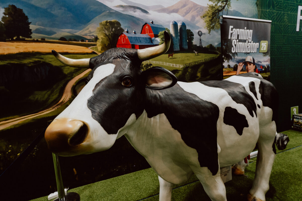 Ein lebensgroßes Modell einer schwarz-weißen Kuh steht in einem Raum auf künstlichem Gras, mit einem Farming Simulator 25-Banner und einer Farmlandschaft im Hintergrund mit Ställen, Silos, Feldern und Bergen.
