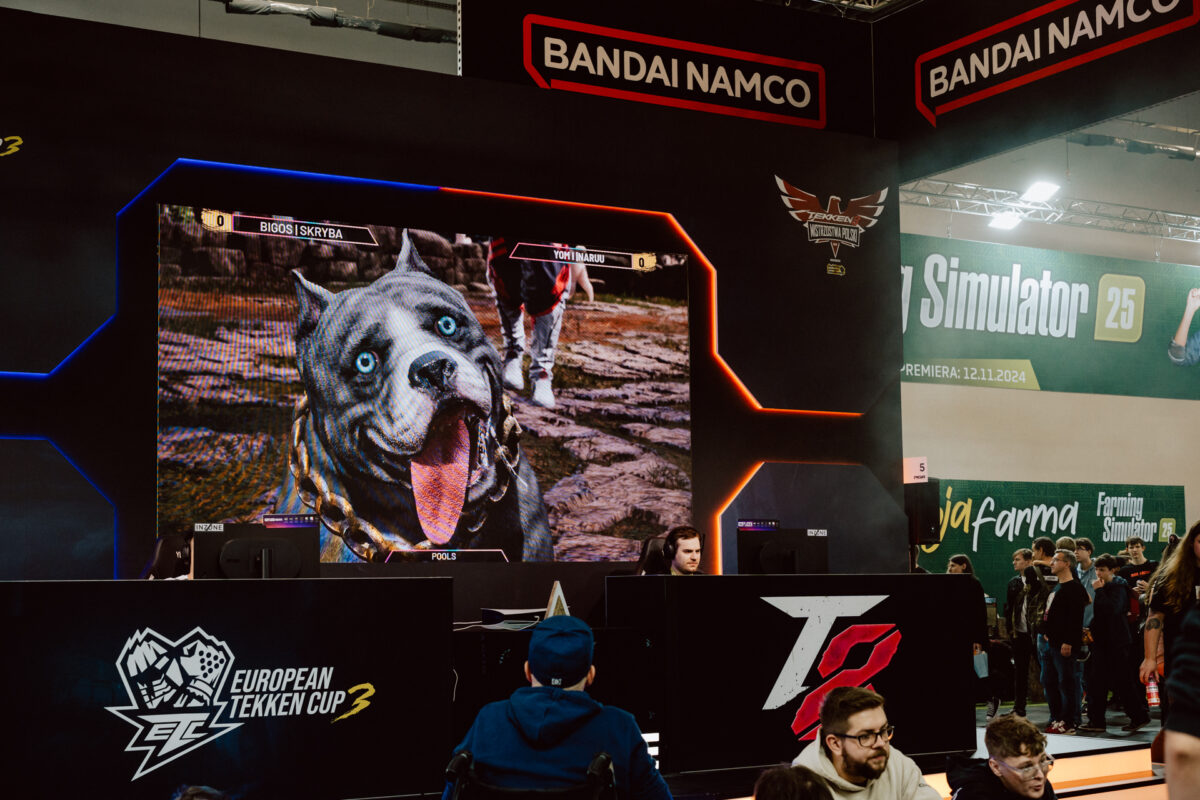 Auf einem großen Bildschirm bei einem Gaming-Event werden Tekken-Spiele mit einer blauäugigen, cartoonartigen Hundefigur gezeigt. Menschen sitzen an Spielstationen, während die Menge zusieht. Bandai Namco und die Veranstaltungsbanner sind oben zu sehen.  