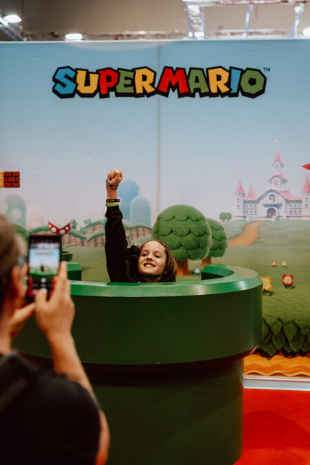 Ein Kind posiert mit erhobenem Arm, der aus einer grünen Röhre vor einem Super-Mario-Hintergrund auftaucht, während jemand ein Foto von ihm macht. Im Hintergrund sind Cartoon-Bäume und ein Schloss zu sehen. 