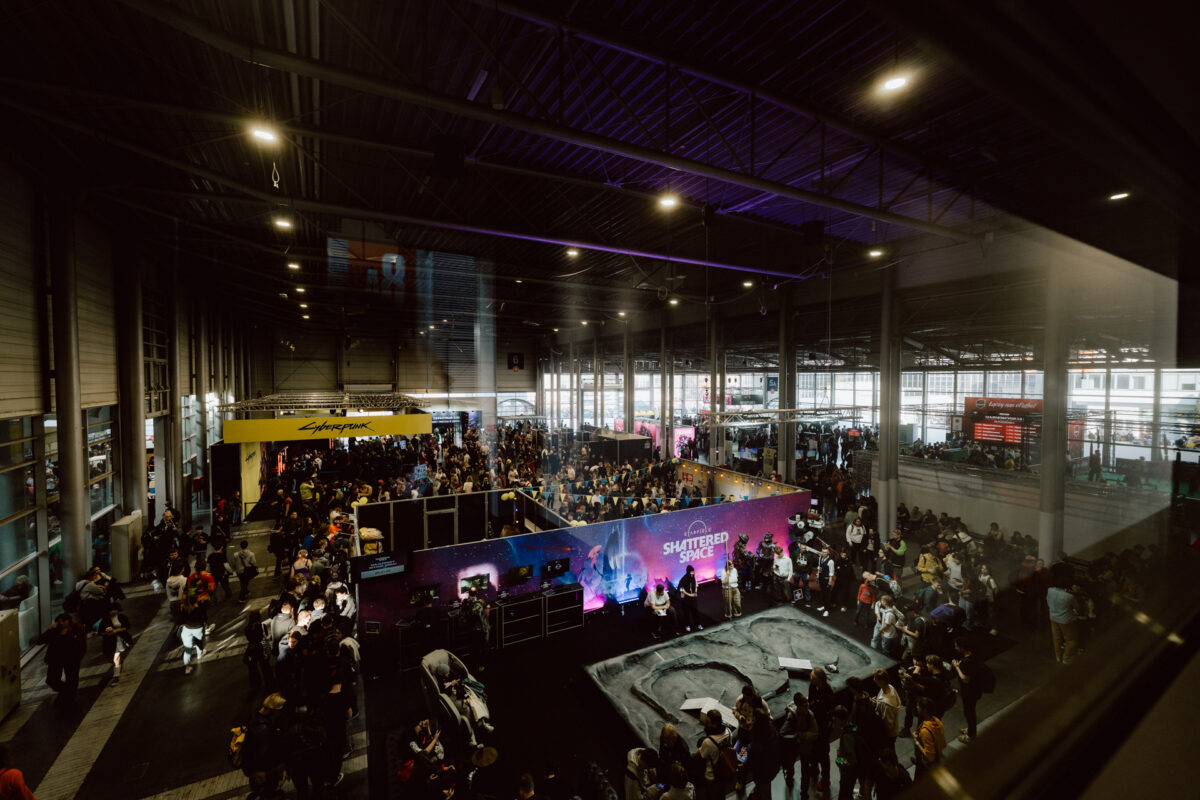 Die große Kongresshalle ist überfüllt mit Besuchern der Veranstaltung. Stände, darunter einer mit der Aufschrift 'Shattered Space', und verschiedene Auslagen sind unter den hellen Lichtern und der hohen industriellen Decke zu sehen. 