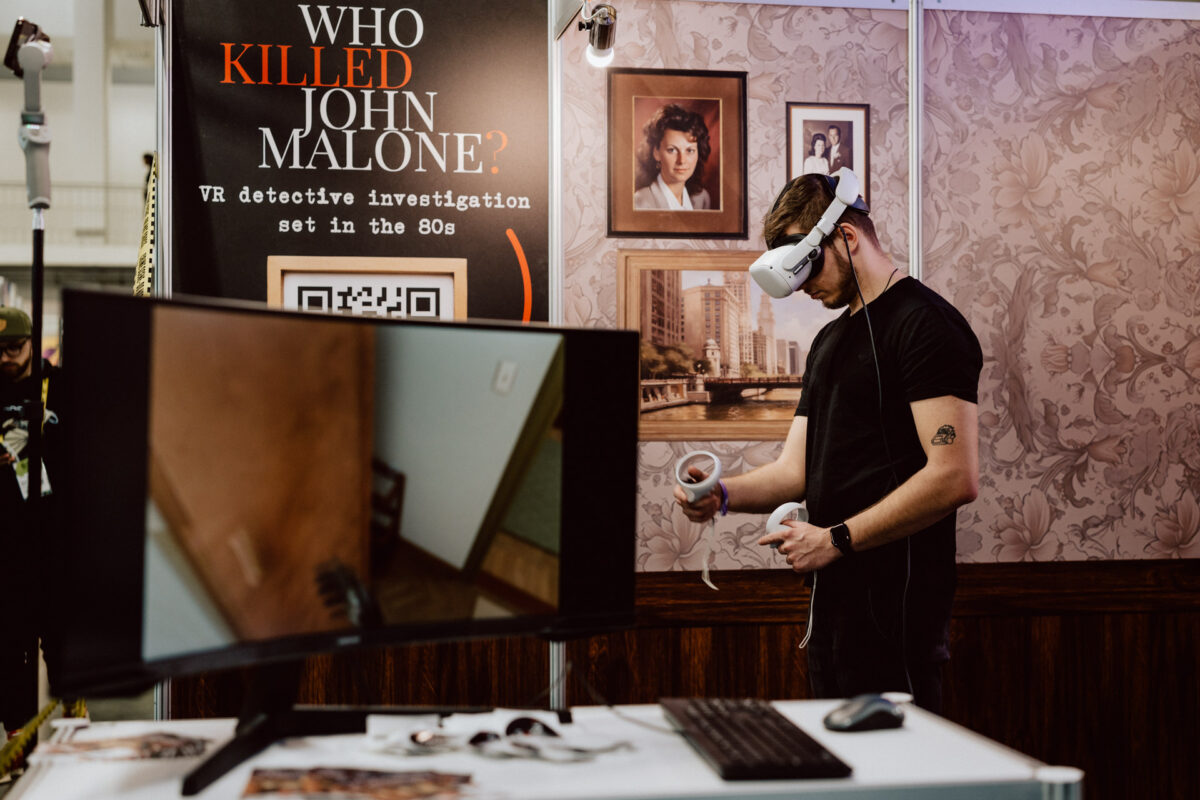 Ein Mann mit einem VR-Headset steht neben einem Computermonitor an einem Ausstellungsstand. Hinter ihm steht ein Schild mit der Aufschrift "Who killed John Malone? Ein VR-Krimi, der in den 1980er Jahren spielt", mit alten Fotos an einer tapezierten Wand. 