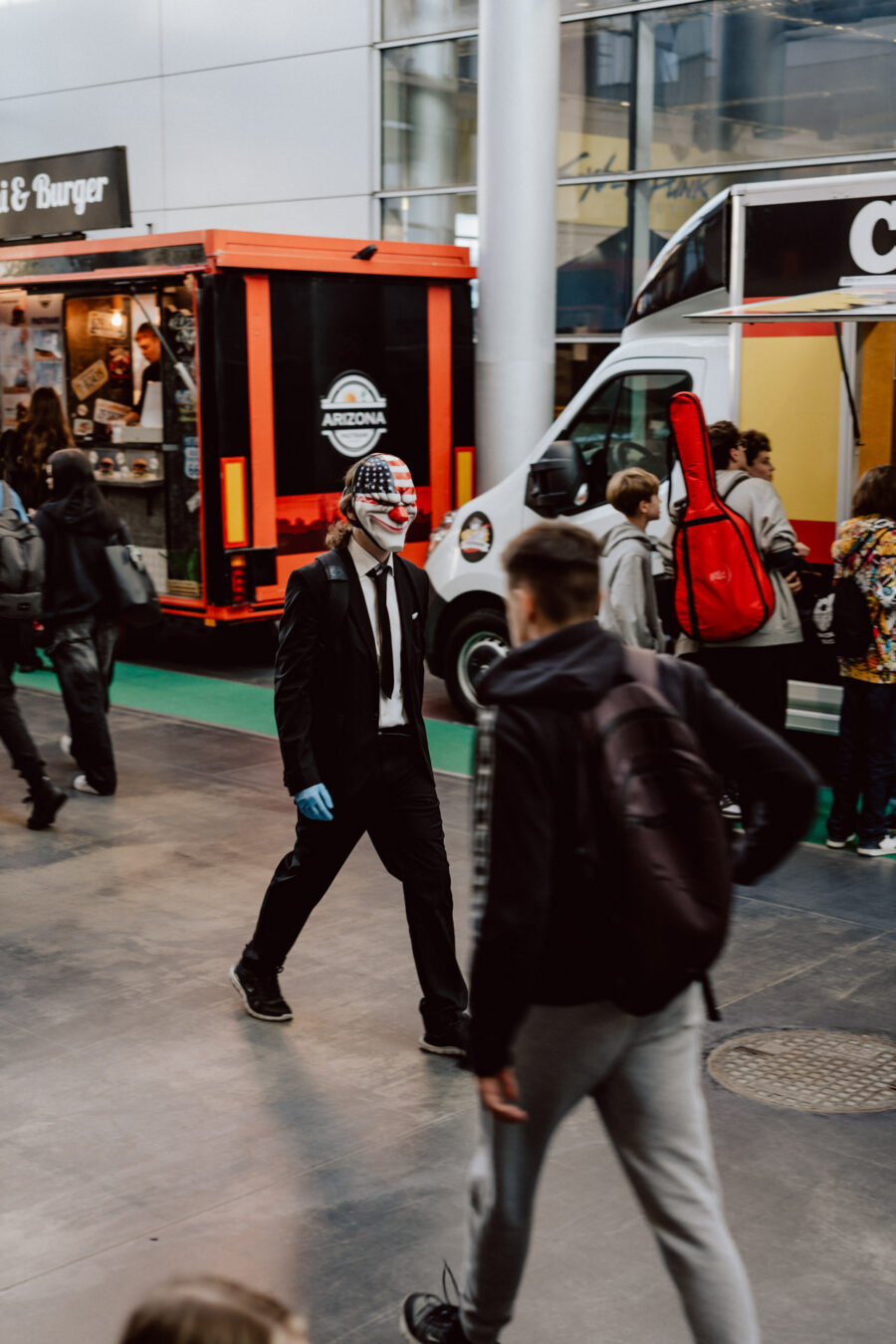 Eine Person im schwarzen Anzug und mit Handschuhen trägt eine Clownsmaske mit einem amerikanischen Flaggenmotiv. Sie geht in der Nähe der Food Trucks spazieren, während andere vorbeigehen und sich im Hintergrund treffen.
