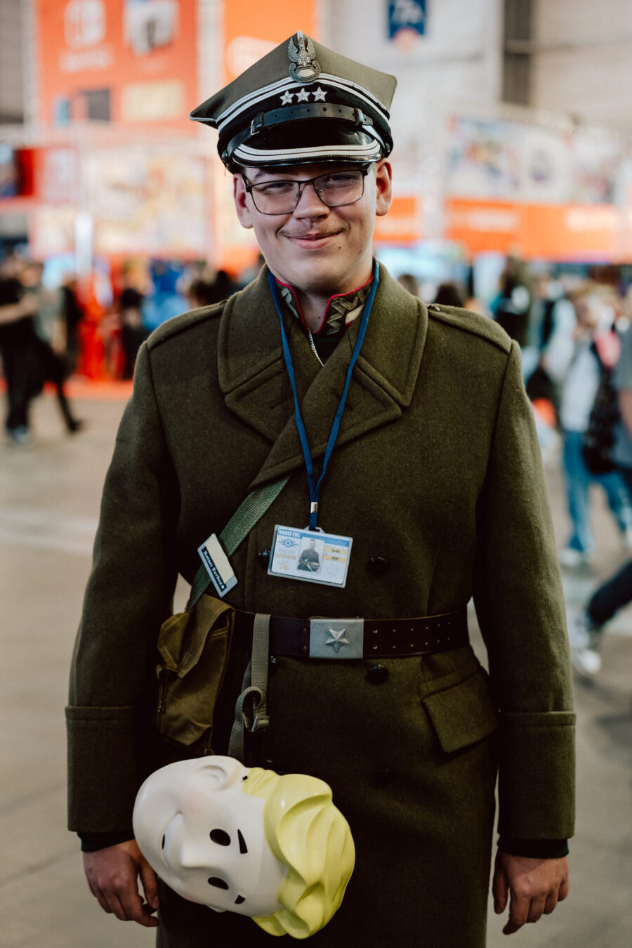 Eine Person in einem Militärmantel und einer Mütze, die eine Brille und einen Ausweis an einem Schlüsselband trägt, steht in einem Raum auf einem Kongress. Er hält eine große Cartoon-Maske mit blondem Haar und einem lächelnden Gesicht in der Hand. Die Kulisse ist mit Menschen und Ständen gefüllt.  