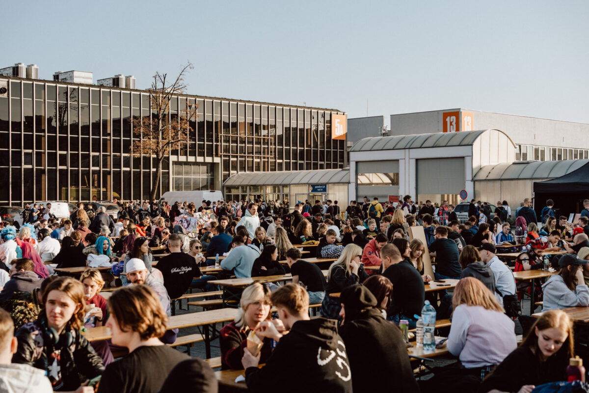 Eine große Menschenmenge sitzt an Holztischen unter freiem Himmel, trifft sich und isst vor den modernen Gebäuden unter freiem Himmel. Die Atmosphäre wirkt lebendig und festlich. 