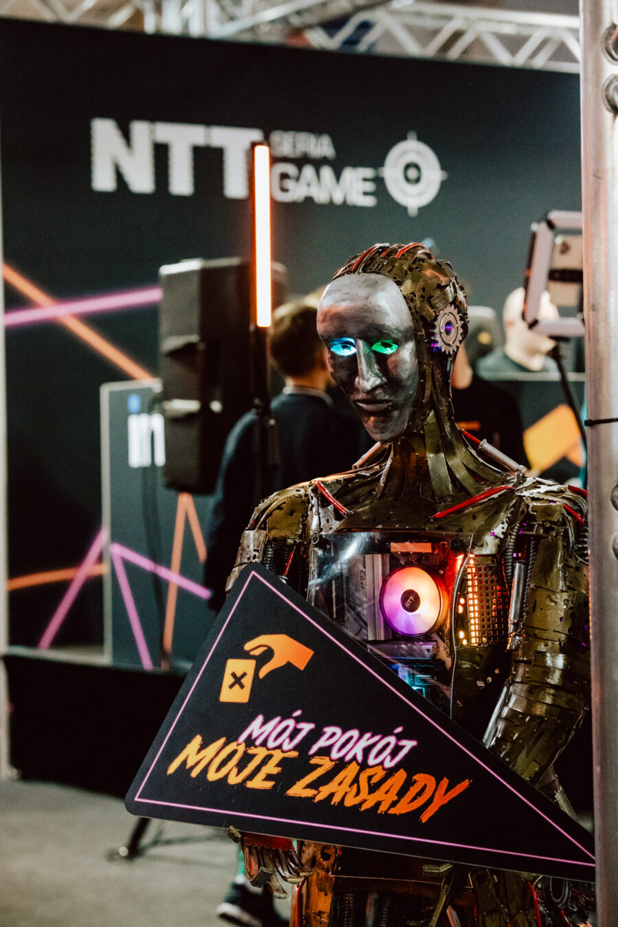 Ein humanoider Roboter, der ein dreieckiges Schild mit polnischem Text hält, steht auf einer Technologieveranstaltung. Im Hintergrund sind unscharfe Menschen und ein 'NTT GAME'-Banner zu sehen.