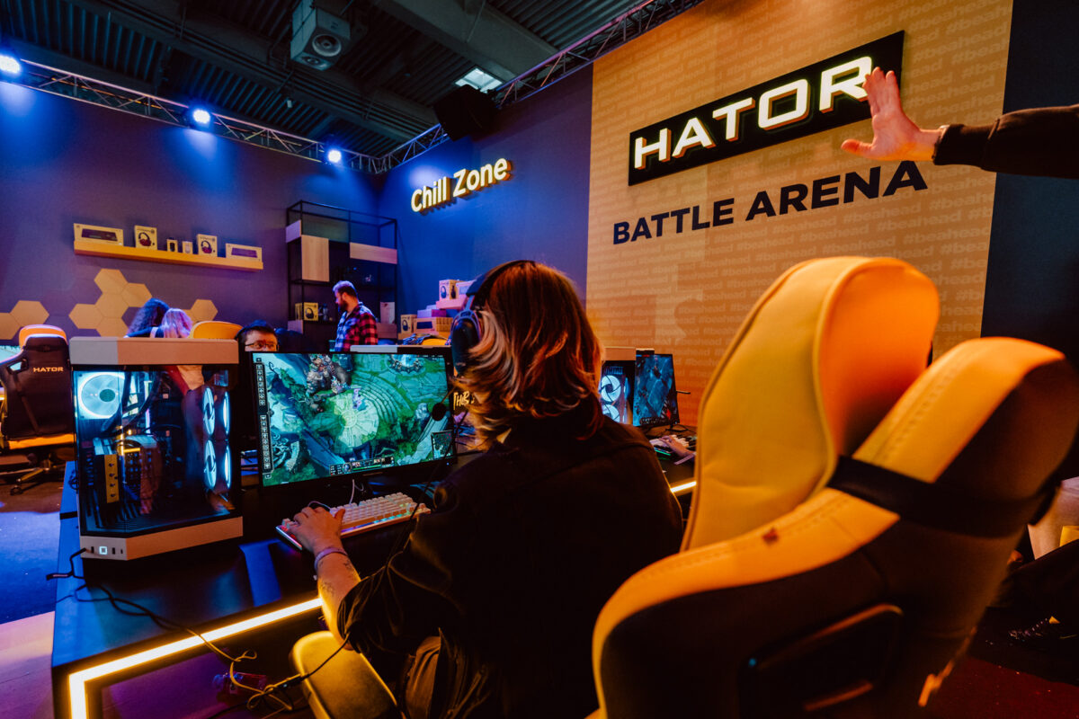 Eine Person mit Kopfhörern spielt ein Computerspiel während eines Gaming-Events in der HATOR Battle Arena. Im Hintergrund sind Gaming-Computer, gelbe Stühle und ein 'Chill Zone'-Schild unter farbiger Beleuchtung zu sehen.