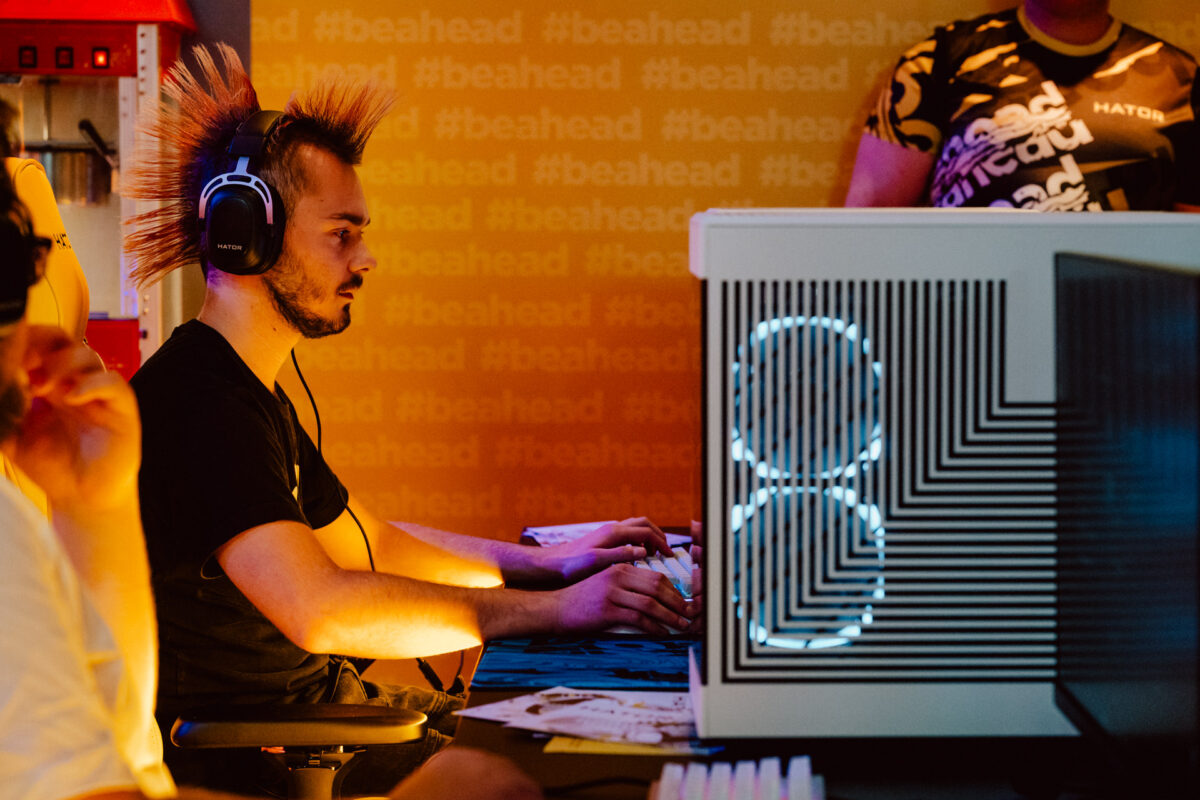 Ein Mann mit einem stacheligen Irokesenschnitt und Kopfhörern sitzt an einem Computer und konzentriert sich auf das Spiel. Der Computer ist von hinten beleuchtet und der gelbe Hintergrund ist '#beahead'. Eine weitere Person steht in der Nähe, teilweise sichtbar. 