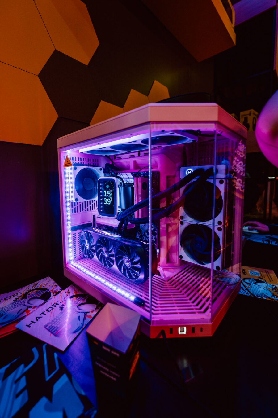 Ein speziell angefertigter Gaming-PC mit internen Komponenten, die von rosa und lila LEDs beleuchtet werden, steht auf einem Schreibtisch neben Zeitschriften und einer kleinen Kiste. Im Inneren sind Kabel, Lüfter und eine große Grafikkarte zu sehen. 