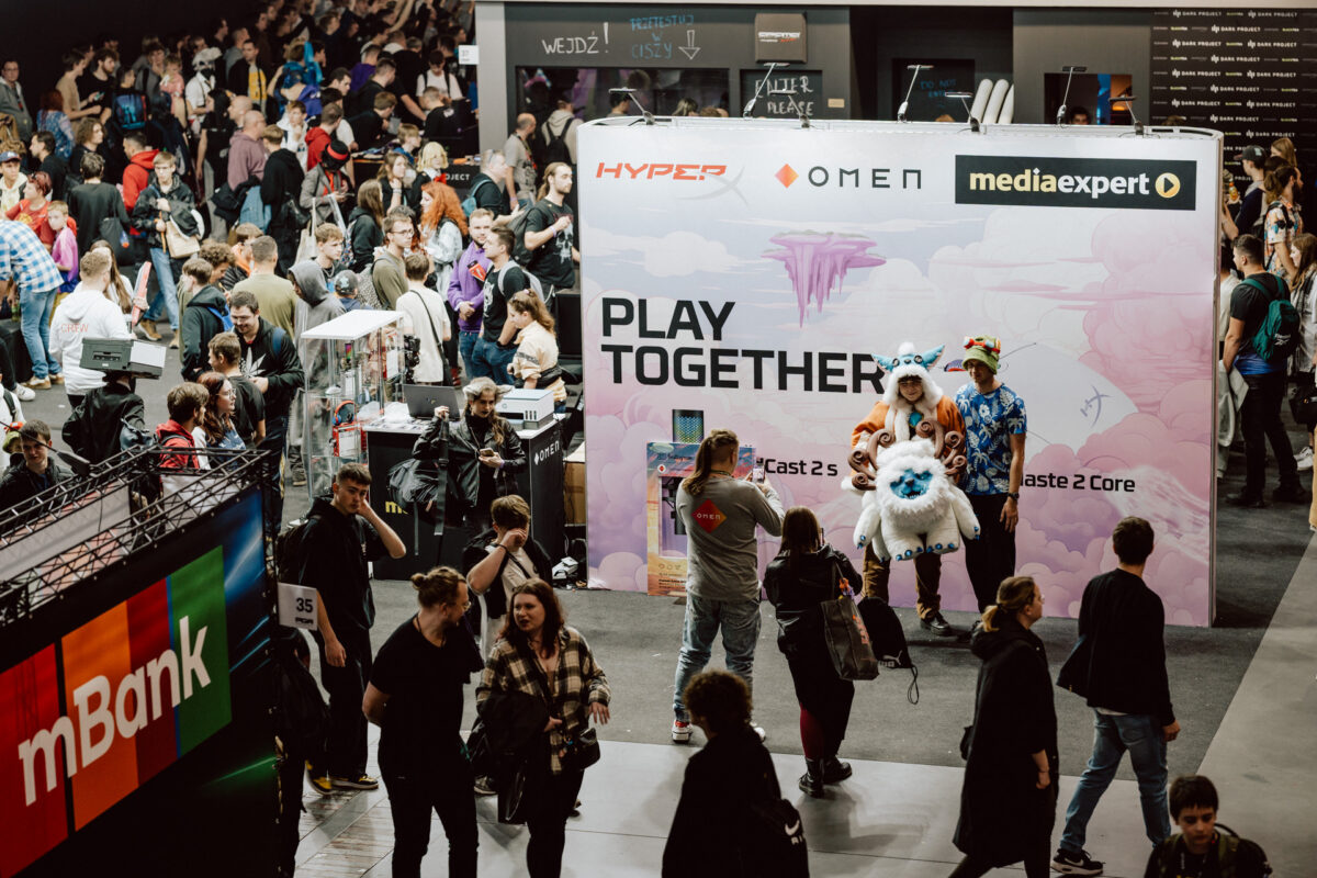 Eine große Menschenmenge nimmt an einer Spieleveranstaltung teil. Zwei Menschen posieren in Kostümen neben dem rosafarbenen 'Play Together'-Stand, während andere in der überfüllten Kongresshalle an den verschiedenen Technologie- und Spieleständen vorbeilaufen oder sich dort versammeln. 