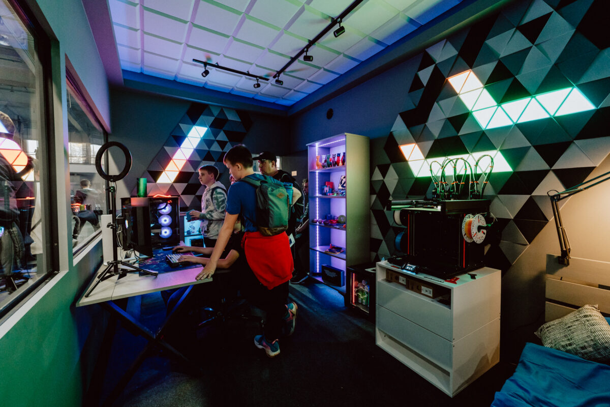 Mehrere Personen stehen um einen Computertisch in einem modernen Raum mit geometrischen Wandpaneelen, bunter LED-Beleuchtung und Regalen mit Elektronik und 3D-Druckern.