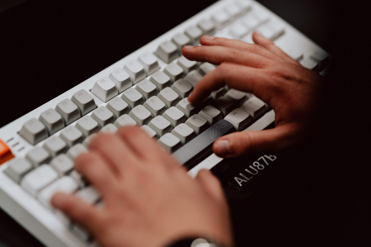 Nahaufnahme von zwei Händen, die auf einer weißen mechanischen Tastatur mit grauen Tasten tippen, von oben gesehen. Die Tastatur hat den Modellcode 'ALU87B', der in der unteren rechten Ecke sichtbar ist. 