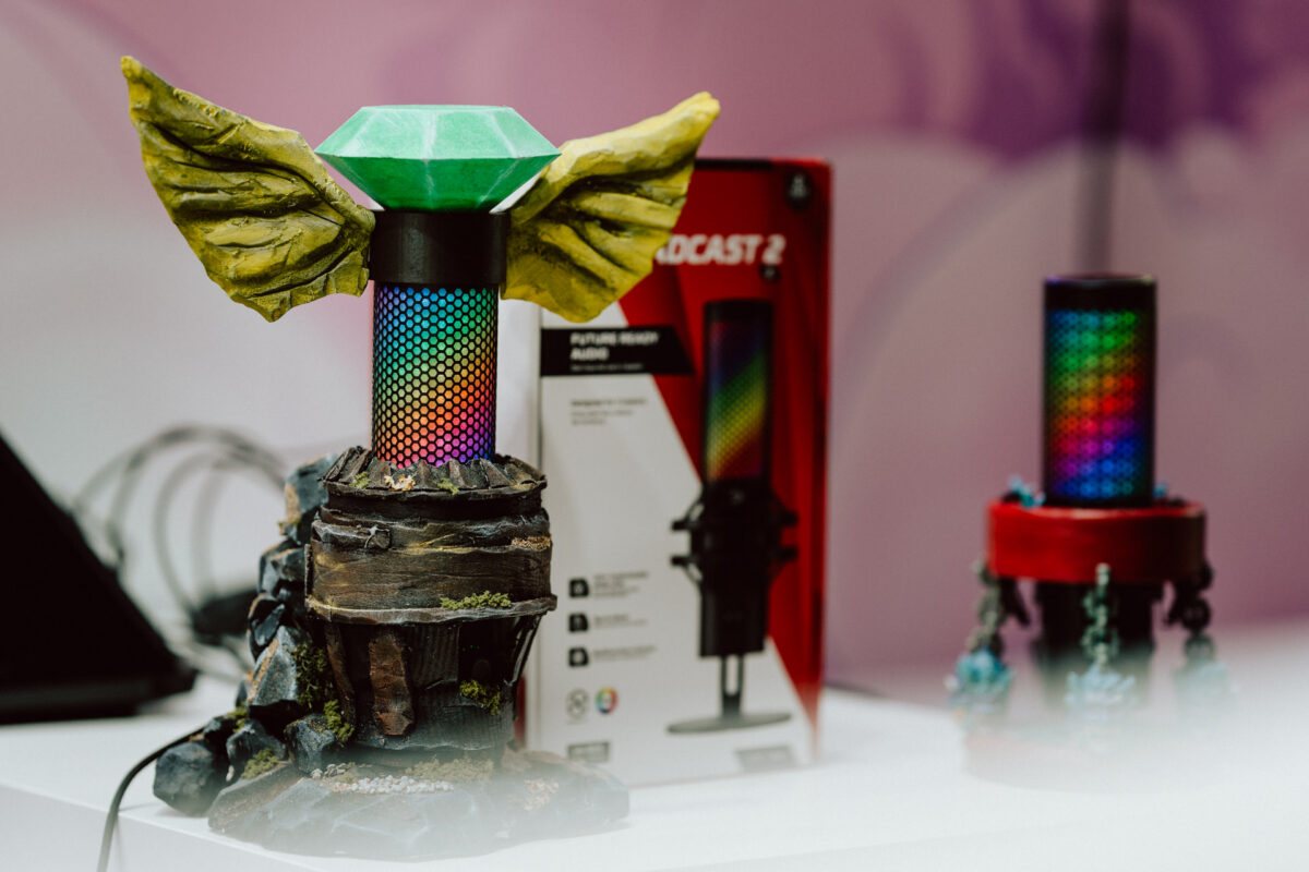 Das farbenfrohe beleuchtete Mikrofon ist mit Fantasyelementen verziert, darunter große gelbe Flügel und ein grüner juwelenartiger Aufsatz. Es wird zusammen mit einer Schachtel und einem weiteren, ebenfalls beleuchteten Mikrofon präsentiert.