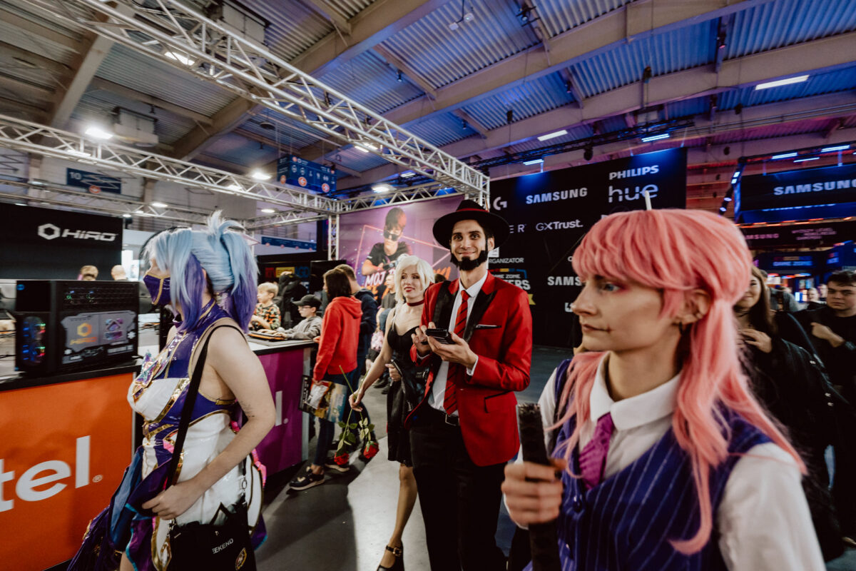 Menschen in farbenfrohen Kostümen laufen durch eine überfüllte Kongresshalle, die mit hellen Lichtern, Technikständen und großen Bannern gefüllt ist. Die Atmosphäre ist lebhaft und energiegeladen, und viele Teilnehmer sind als Kostüme verkleidet. 