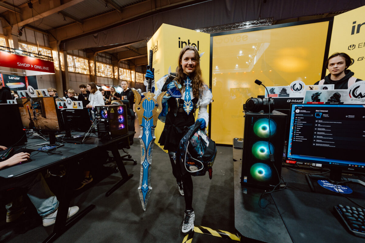 Ein Cosplayer in einem blau-weißen Outfit, der ein großes, verziertes Schwert in der Hand hält, steht vor einem gelben Intel-Stand auf einem Gaming-Event. Im Hintergrund sind Computer und andere Teilnehmer zu sehen.