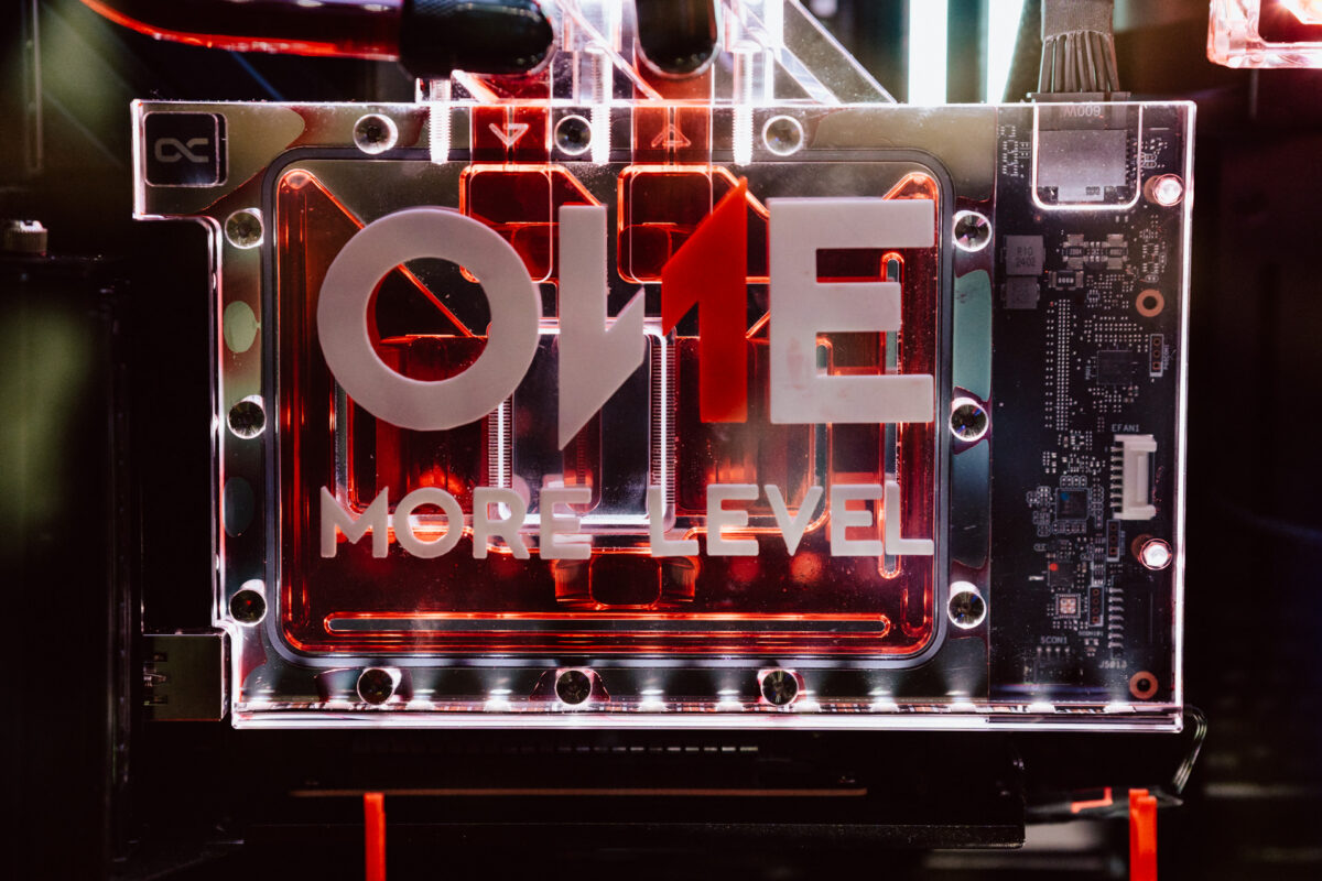Nahaufnahme von hintergrundbeleuchteter Computer-Hardware mit roten Flüssigkühlungsröhren und der Aufschrift 'ONE MORE LEVEL' auf einer transparenten Platte. Das Bild zeigt einen Teil der Platine auf der rechten Seite. 