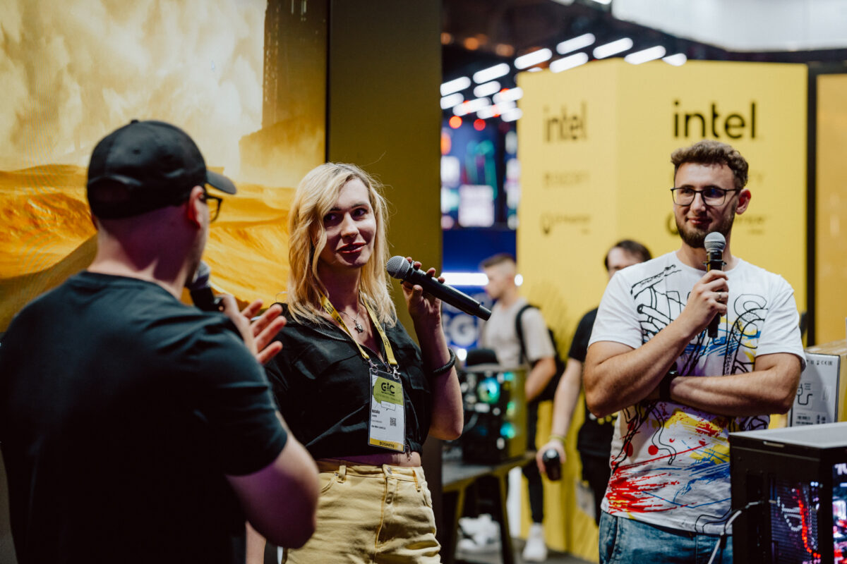 Drei Personen stehen in einem Raum bei einer Intel-Veranstaltung, zwei halten Mikrofone und eine gestikuliert, während sie spricht. Im Hintergrund sehen Sie gelbe Intel-Banner und Computerausrüstung. Die Atmosphäre ist lebhaft und einnehmend.  