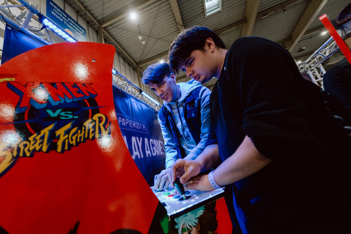 Zwei Personen spielen eifrig das Arcade-Spiel X-Men vs. Street Fighter auf einer Indoor-Party. Sie konzentrieren sich auf die Steuerung und den Bildschirm, umgeben von hellen Lichtern und Werbebannern. 