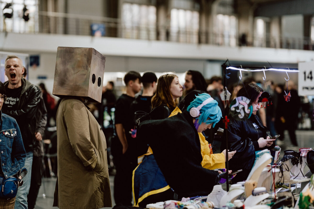 Eine geschäftige Kongressszene mit verkleideten Menschen, darunter eine Person mit einer Schachtel auf dem Kopf und eine andere mit einer blauen Perücke und Kopfhörern an einem Stand. Die Besucher begutachten die Tische mit den Waren und Auslagen in der belebten Halle. 