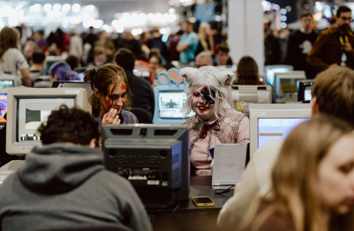 Menschen in Kostümen und mit bemalten Gesichtern sitzen bei einer überfüllten Veranstaltung an Computerstationen im Retro-Stil. Die Kulisse ist mit Teilnehmern gefüllt, die eine lebendige und pulsierende Atmosphäre schaffen. 