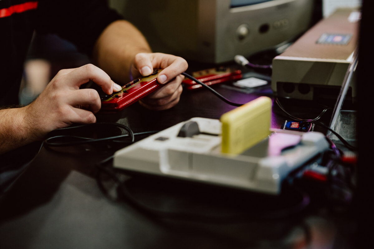 Nahaufnahme einer Person, die einen Spiele-Controller in der Hand hält und eine Retro-Spielkonsole mit einer gelben Kassette und anderer Vintage-Elektronik auf dem Tisch spielt.
