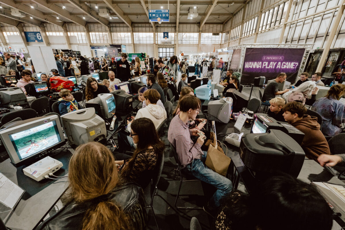 Eine überfüllte Spieleveranstaltung in einer großen Halle mit vielen Menschen, die Videospiele auf alten Röhrenmonitoren spielen; Gruppen und Einzelpersonen konzentrieren sich auf ihre Bildschirme, umgeben von geschäftigem Treiben und Ständen im Hintergrund.