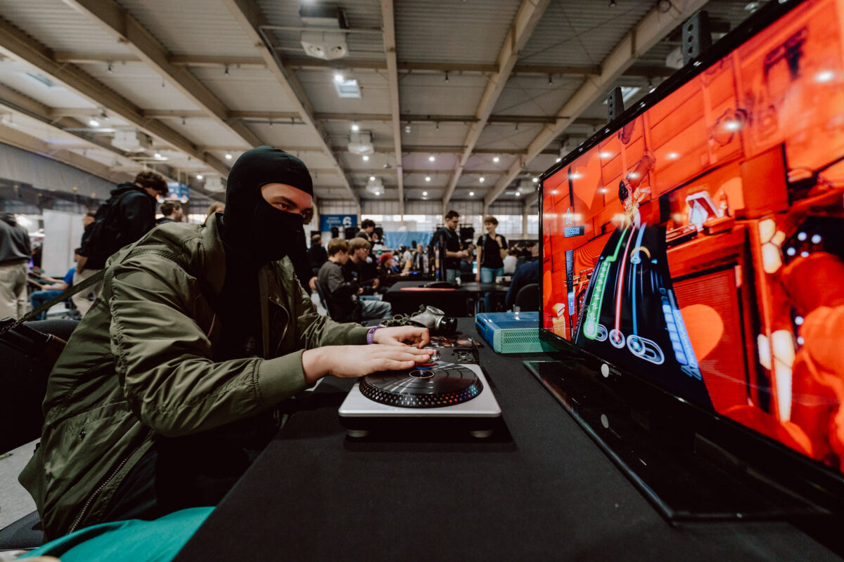 Eine Person, die einen schwarzen Mantel und eine grüne Jacke trägt, spielt während eines Gaming-Events eine Simulation eines DJs auf einem Plattenspieler-Controller, wobei das bunte Spiel auf einem großen Monitor angezeigt wird. Andere Teilnehmer sind im Hintergrund zu sehen. 