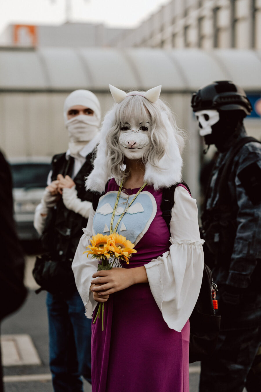 Eine Person mit einer weißen Tiermaske, einer langen weißen Perücke und einem lila Kleid, die gelbe Blumen hält, steht draußen. Zwei Personen in dunklen taktischen Outfits und Masken stehen in der Nähe. Im Hintergrund ist ein Gebäude mit Glasfenstern zu sehen.  