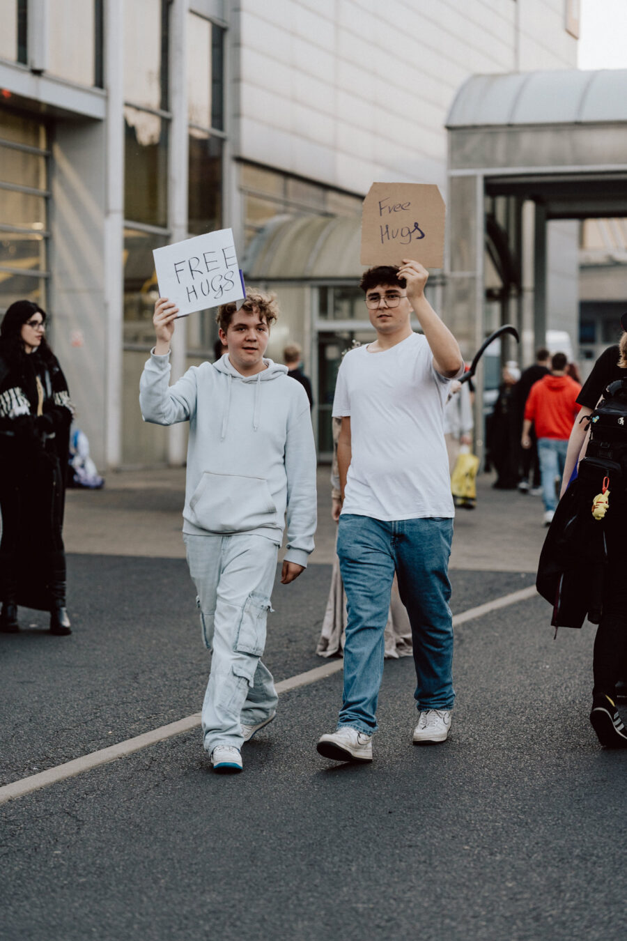 Zwei junge Männer gehen eine Straße in der Stadt entlang und halten Plakate mit der Aufschrift 'FREE HUGS'. Sie sind umgeben von anderen Menschen, einige gehen, andere stehen, in der Nähe eines modernen Gebäudes. 