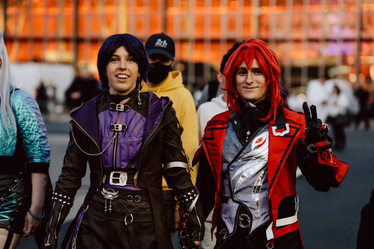 Zwei Personen in farbenfrohen Cosplay-Kostümen, eine mit kurzen blauen Haaren in Lila und Schwarz, die andere mit langen roten Haaren in Rot und Schwarz, stehen draußen und lächeln, mit der Menge und einem orange beleuchteten Gebäude im Hintergrund.