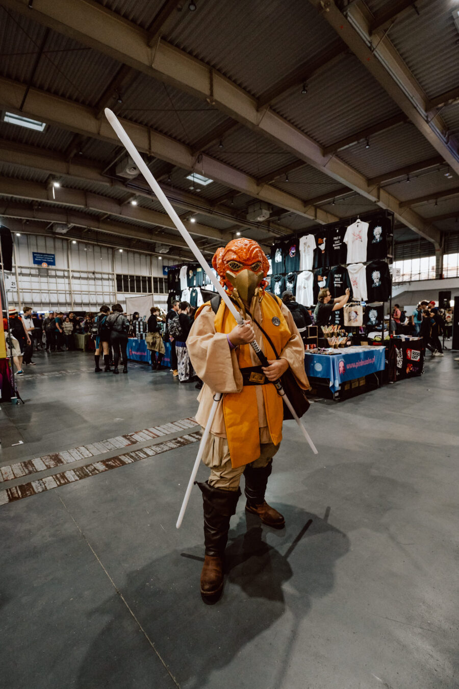 Eine Person mit einer orangefarbenen Maske und einer Jedi-Robe posiert mit einem langen weißen Stock auf einem Kongress, mit Verkaufsständen und anderen Teilnehmern im Hintergrund in einer großen Halle.