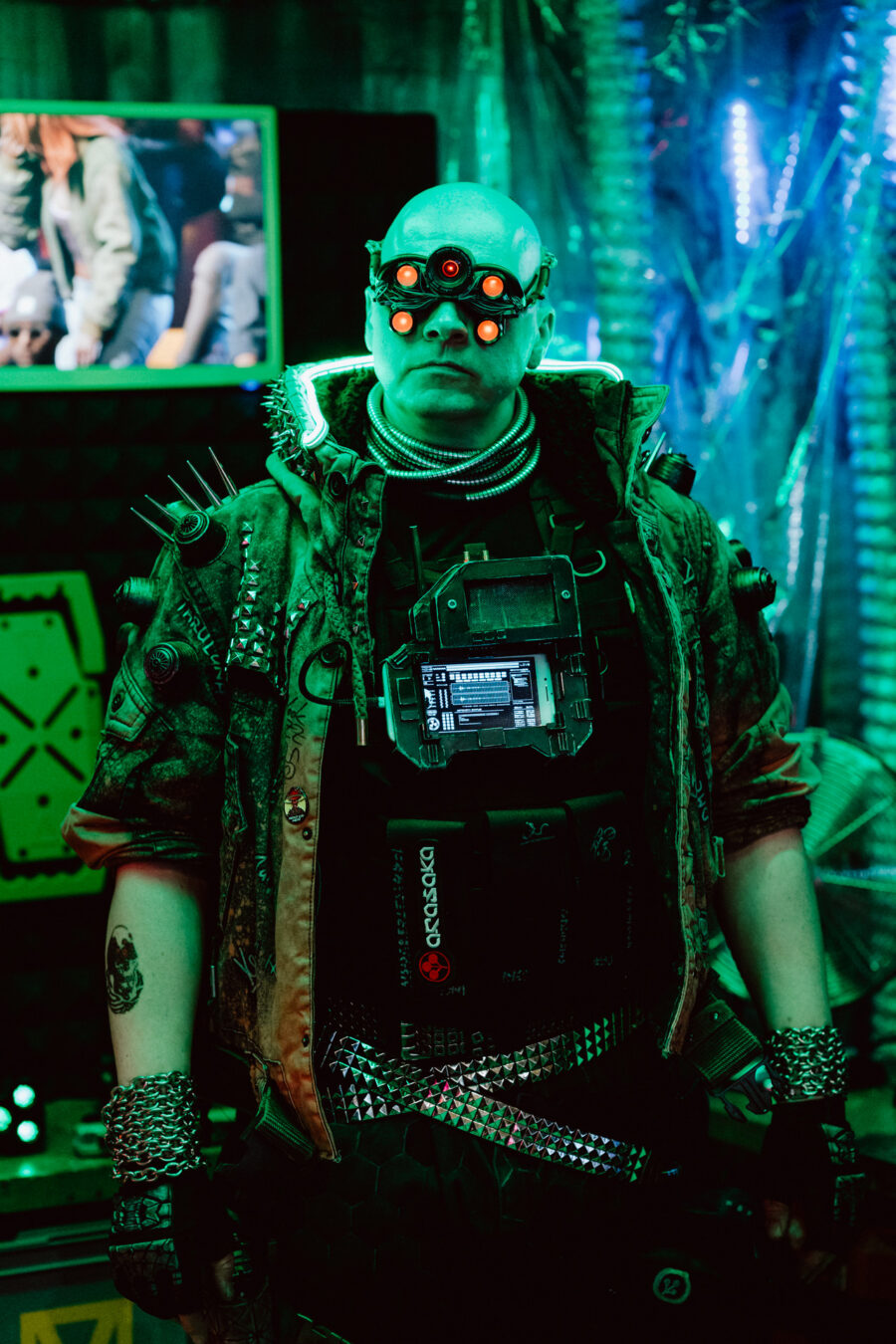 Eine Person in einem futuristischen Cyberpunk-Outfit mit leuchtenden Brillen, aufgespießten Epauletten und elektronischen Geräten auf der Brust steht in einer neonfarbenen High-Tech-Umgebung.