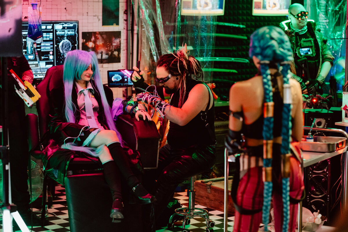 Eine Person mit blauer Perücke und Kostüm sitzt auf einem Stuhl, während eine andere Person mit Dreadlocks und Schutzbrille in einem neonbeleuchteten, futuristischen Raum mit Cyberpunk-Dekor und verschiedenen Geräten an einem Arm arbeitet.