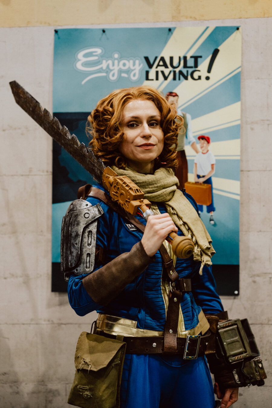 Eine Person in einem blauen Overall, Rüstung und Schal hält ein großes Schwert vor einem Plakat mit der Aufschrift "Enjoy Vault Living!". Das Kostüm erinnert an eine Figur aus dem Spiel Fallout auf einer Convention oder einem Cosplay-Event. 