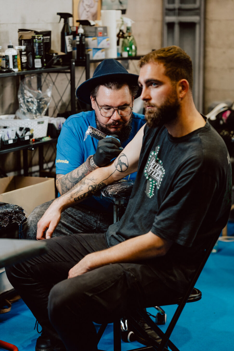 Ein Tätowierer, der Handschuhe und eine Mütze trägt, tätowiert in einem Studio den Unterarm eines Mannes. Der Mann sitzt ruhig da und schaut in die Ferne, während der Künstler sich auf seine Arbeit konzentriert. Im Hintergrund sind Tattoo-Ausrüstung und Zubehör zu sehen.  