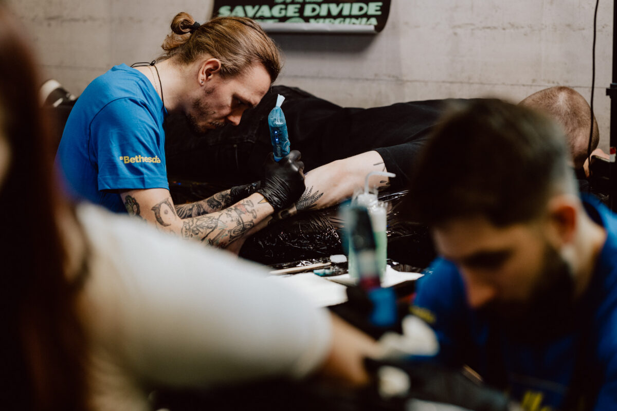 Ein Tätowierer mit Tätowierungen auf den Armen konzentriert sich auf die Tätowierung des Beins des Kunden; ein anderer Künstler arbeitet im Vordergrund, während die Tätowiermaterialien auf dem Tisch zu sehen sind.