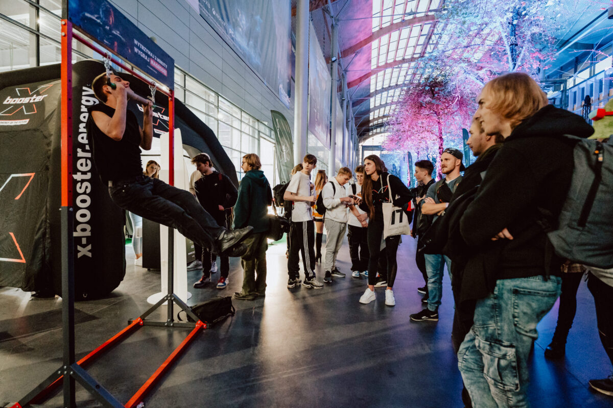 Ein Mann zieht an einer Bar im Raum, während eine Gruppe von Leuten zusieht. Der Raum ist modern, mit hohen Decken und farbenfroher Beleuchtung und Werbebannern und Ständern im Hintergrund. 