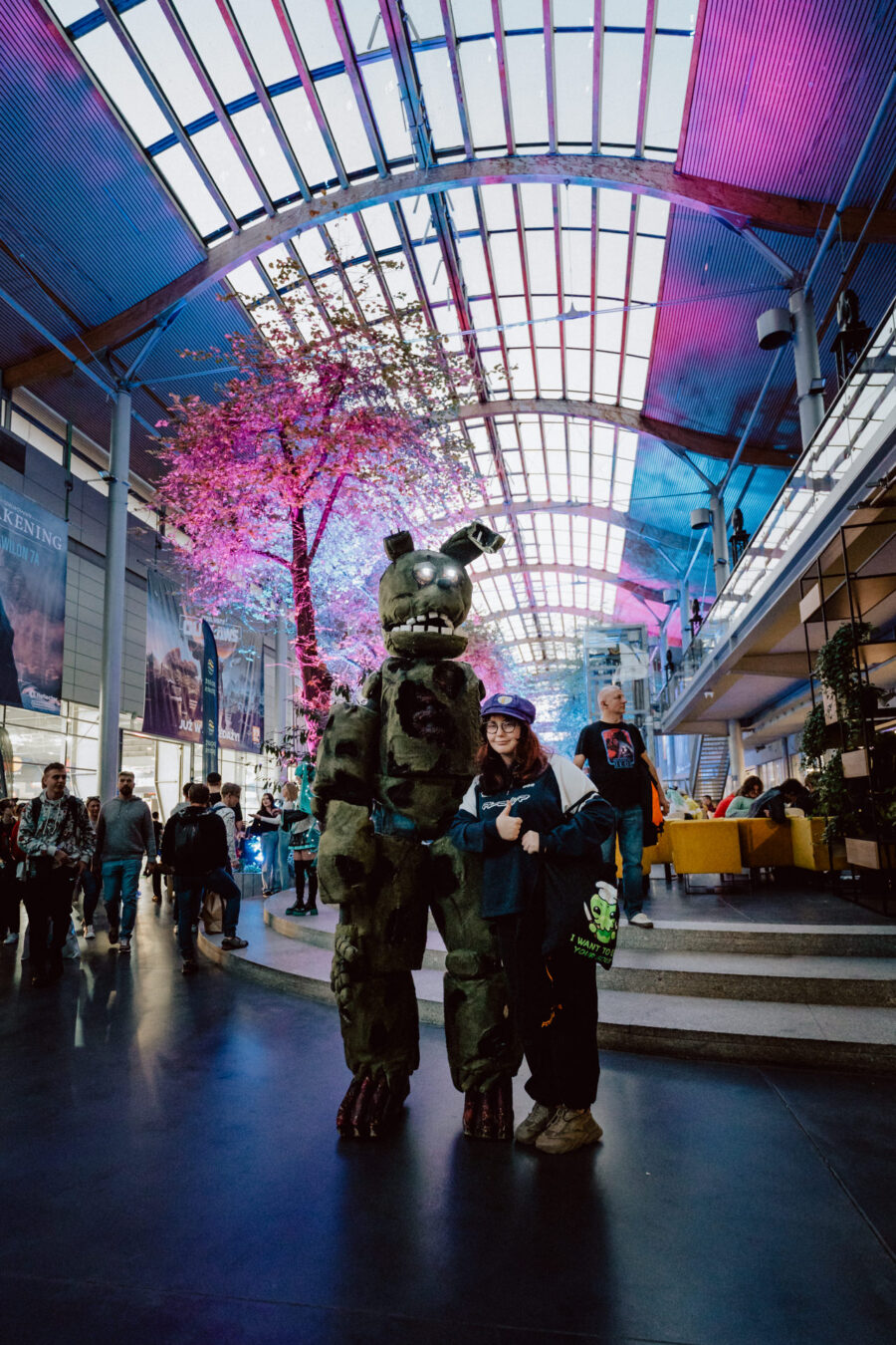 Eine Person in einem riesigen grünen animatronischen Kaninchenkostüm posiert mit einem lächelnden Teilnehmer in einem geräumigen, modernen Kongresszentrum mit einer Glasdecke und einem verschnörkelten rosa Baum. Im Hintergrund flanieren andere Menschen und versammeln sich. 