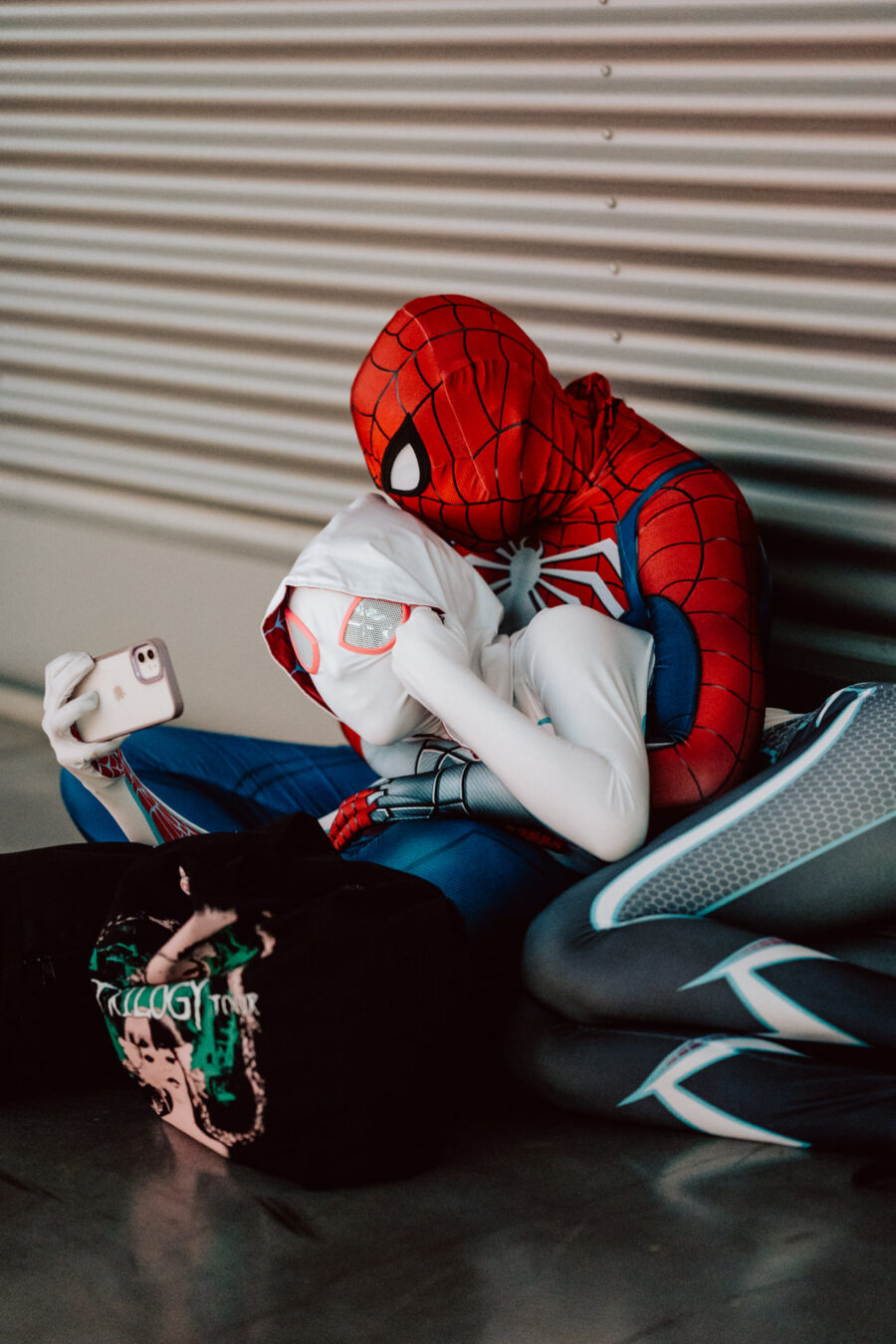Zwei Personen in Spider-Man-Kostümen sitzen auf dem Boden, einer hält ein Selfie-Handy in der Hand, der andere stützt seinen Kopf auf seine Schulter. In der Nähe liegt ein gemusterter Rucksack und im Hintergrund ist eine Metallwand zu sehen. 