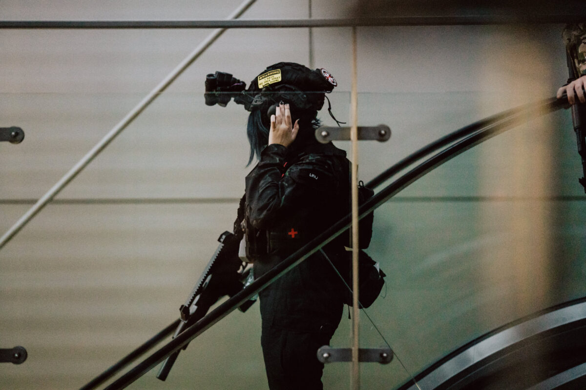 Eine Person in taktischer Kleidung und mit Helm, die ein Gewehr in der Hand hält, steht auf einer Rolltreppe in einem Gebäude, seitwärts gedreht, und berührt den Helm mit einer Hand.