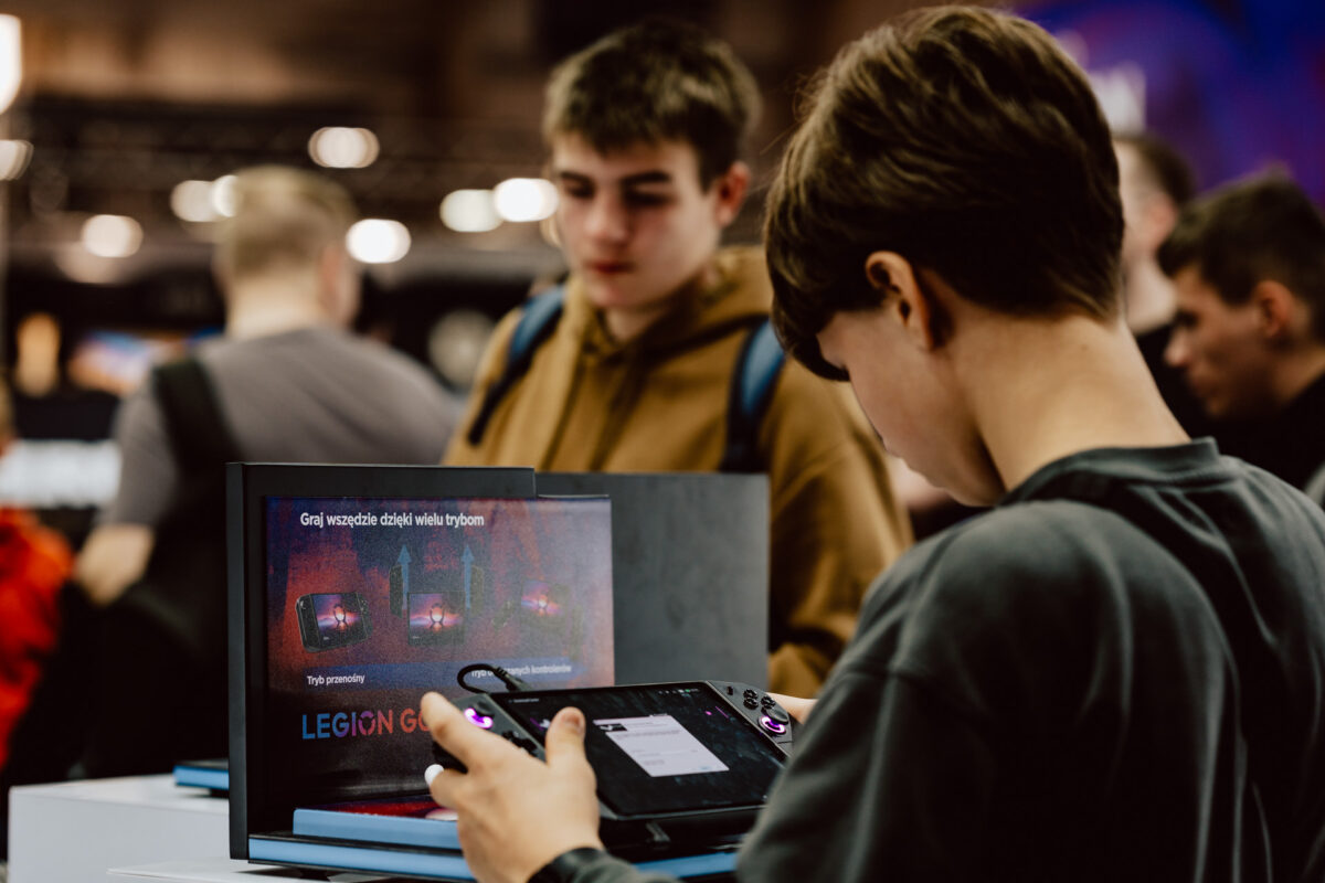 Ein Junge mit Brille benutzt ein tragbares Spielgerät bei einer Technologieveranstaltung. Im Hintergrund sind andere Teilnehmer und Spielbildschirme zu sehen.